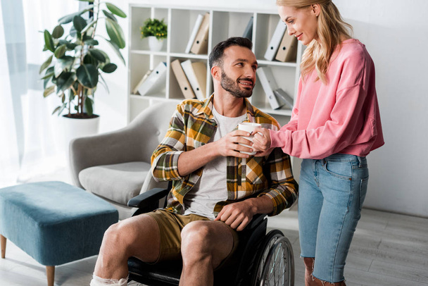 cheerful woman giving cup of tea to bearded man in wheelchair  - Foto, immagini