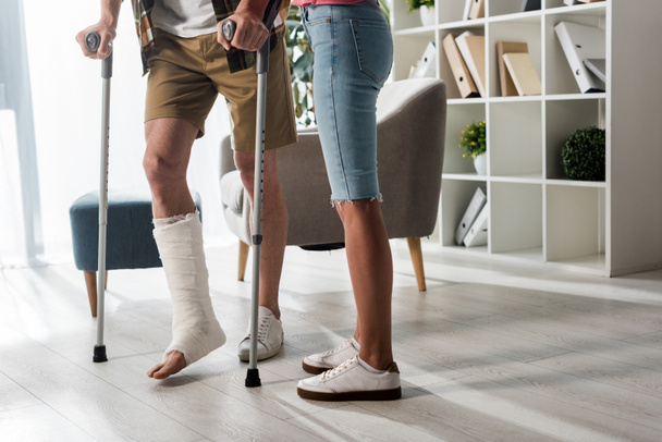 cropped view of woman standing near man holding crutches at home  - Фото, изображение