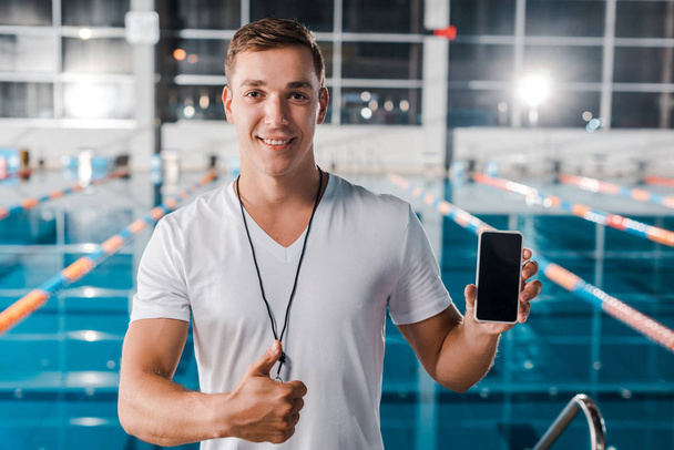 boldog edző mutatja hüvelykujját fel, miközben gazdaság smartphone üres képernyőn  - Fotó, kép