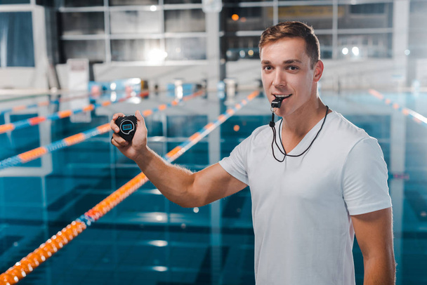 smiling trainer with timer in hand holding whistle in mouth  - Fotoğraf, Görsel