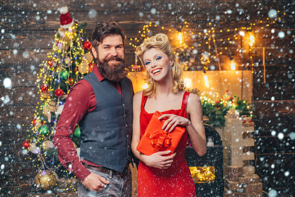 Sexy couple with gift over Christmas tree background. Portrait of a beautiful couple in Christmas dress holding gift while standing and looking up over Christmas tree background. - Fotó, kép