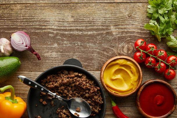 Vista superior de la carne picada en sartén con salsas y verduras crudas sobre fondo de madera
 - Foto, Imagen