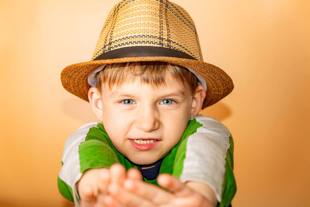 Ένα αγόρι με ψάθινο καπέλο και πράσινα ρούχα σε μπεζ φόντο βγάζει το καπέλο του χαιρετώντας. - Φωτογραφία, εικόνα