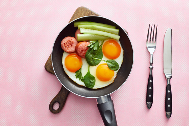 top view of fried eggs with spinach leaves, cucumber and sausage in frying pan near cutlery on pink dotted background - Photo, Image