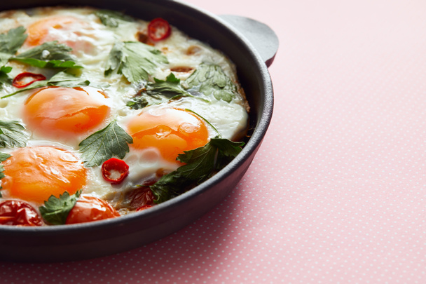 close up view of fried eggs with parsley, tomatoes and chili pepper in frying pan on pink background - Fotoğraf, Görsel