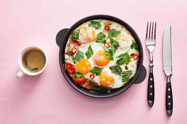 top view of fried eggs with parsley, tomatoes and chili pepper in frying pan near cutlery and coffee on pink background - Photo, Image