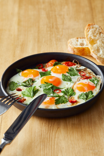 fried eggs with parsley and chili pepper on wooden table with cutlery and bread - Photo, image