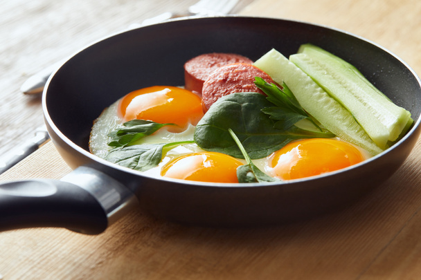 fried eggs in frying pan with spinach, cucumber and sausage at wooden table - Foto, Bild
