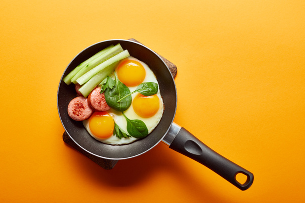 top view of fried eggs with spinach leaves, cucumber and sausage in frying pan on orange background - Foto, Imagem
