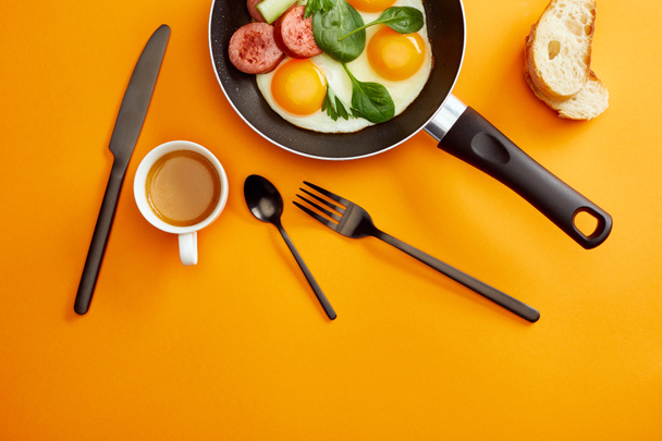top view of fried eggs with spinach leaves, cucumber and sausage in frying pan near coffee, cutlery and bread on orange background - Zdjęcie, obraz
