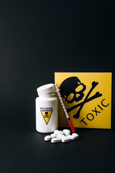 Jar with poison signs by pills and syringe isolated on black - 写真・画像