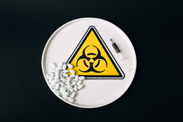 Top view of pills and syringe on plate with biohazard sign isolated on black - Fotografie, Obrázek