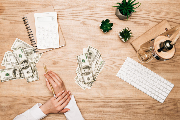 Top view of businesswoman holding pencil near dollar banknotes, plants and computer keyboard  - Фото, изображение