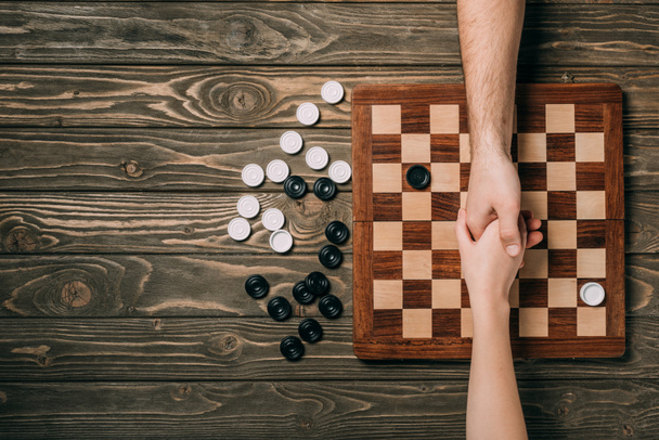 Cropped view of man and woman shaking hands above checkerboard with checkers on wooden background - Fotoğraf, Görsel