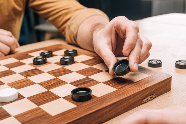 Selective focus of man holding black checker on wooden checkerboard - Foto, afbeelding