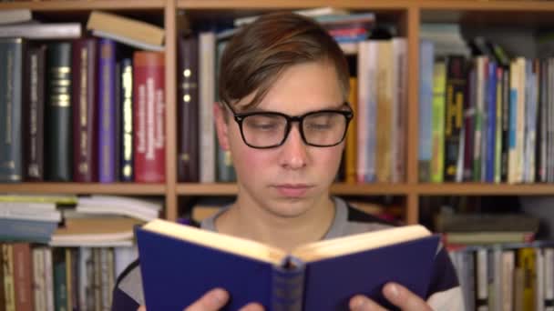 Ένας νεαρός διαβάζει ένα βιβλίο και το χτυπάει απότομα στη βιβλιοθήκη. Ένας άντρας με γυαλιά κοιτάζει προσεκτικά το βιβλίο και κοιτάζει την κάμερα. Στο βάθος υπάρχουν βιβλία στα ράφια των βιβλιοθηκών. Βιβλιοθήκη βιβλίων. - Πλάνα, βίντεο
