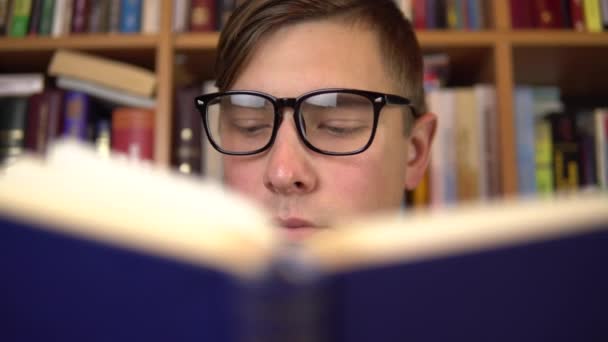Een jongeman leest een boek in een bibliotheek. Een man met een bril kijkt zorgvuldig naar het boek van dichtbij. Op de achtergrond staan boeken op boekenplanken. Boekenbibliotheek. - Video