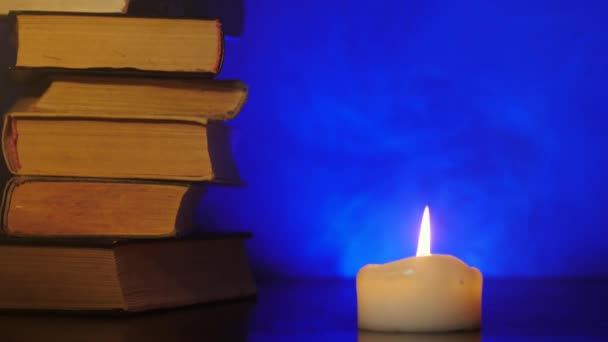 Кучи книг стоят возле горящей свечи и цветной волшебный дым летит
. - Кадры, видео