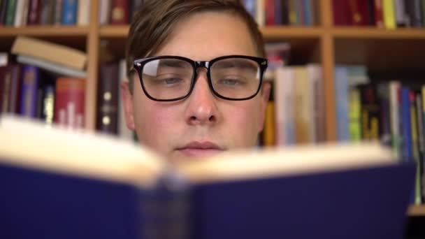 Een jongeman leest een boek in een bibliotheek. Een man met een bril kijkt zorgvuldig naar het boek van dichtbij. Op de achtergrond staan boeken op boekenplanken. Boekenbibliotheek. - Video