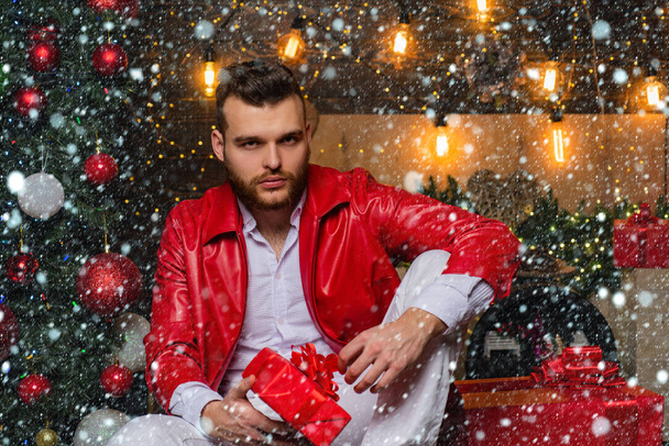 Sexy hot boy holding christmas present in his hand and wearing red leather jacket. Bearded handsome man at full christmas decorated background. Christmas tree, lights and gifts concept. - Foto, Imagem