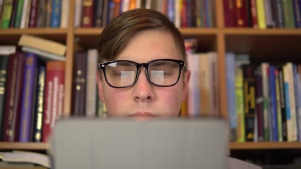 A young man is reading a book in a tablet. A man with glasses carefully looks at the tablet. In the background are books on bookshelves. Book library. - Footage, Video