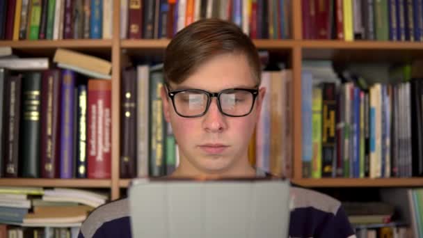 Mladý muž čte knihu v tabletu. Muž s brýlemi si tabletu pozorně prohlíží. V pozadí jsou knihy na regálech. Knihovna knih. - Záběry, video