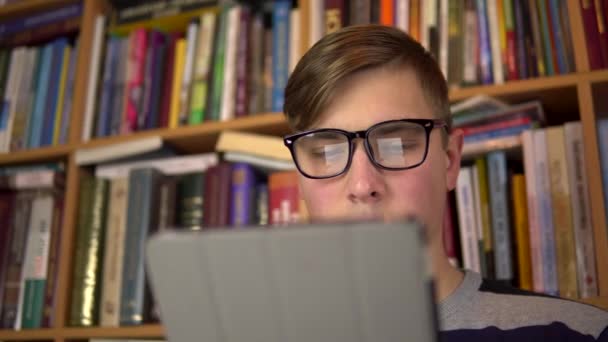 Молодий чоловік читає книжку в таблетці. Чоловік в окулярах уважно дивиться на планшет. На задньому плані знаходяться книги на книжкових полицях. бібліотека книг
. - Кадри, відео
