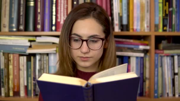 Молода жінка читає книжку в бібліотеці. Жінка в окулярах ретельно дивиться на книгу крупним планом. На задньому плані знаходяться книги на книжкових полицях. бібліотека книг
. - Кадри, відео