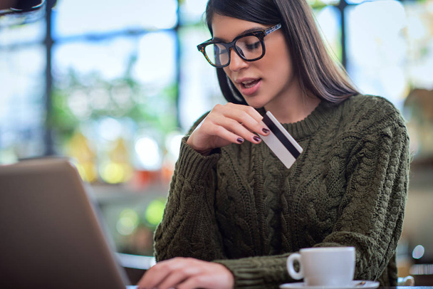 Atractiva morena caucásica en suéter sentada en la cafetería, tecleando en el teclado y sosteniendo la tarjeta de crédito
. - Foto, imagen