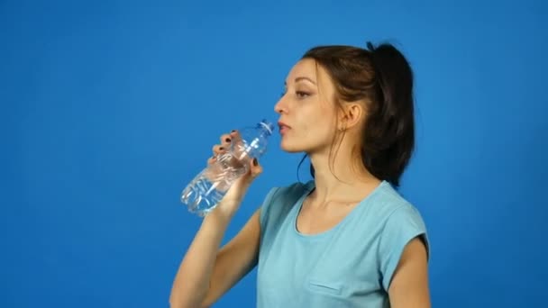 Seitenansicht des brünetten Mädchens mit langen dunklen Haaren trinkt nach dem Training auf blauem Hintergrund im Studio reines Wasser aus einer durchsichtigen Plastikflasche - Filmmaterial, Video