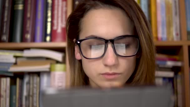 Egy fiatal nő könyvet olvas egy táblagépben. Egy szemüveges nő alaposan megnézi a tablettát. A háttérben könyvespolcokról szóló könyvek vannak. Könyvtár. - Felvétel, videó