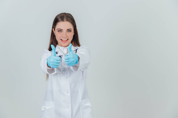 Εμπνευσμένη νεαρή γυναίκα ιατρός με μεγάλα μπλε μάτια, στέκεται, φοράει ιατρική στολή, στηθοσκόπιο πάνω από το λαιμό, χαμογελώντας στην κάμερα, δίνοντας αντίχειρες επάνω. - Φωτογραφία, εικόνα