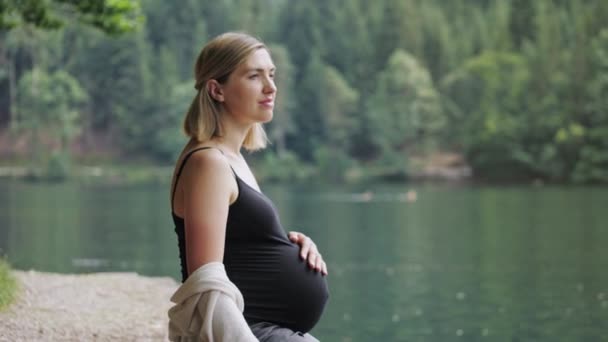 Έγκυος γυναίκα αγγίζει απαλά την κοιλιά της όταν κάθεται κοντά στο βουνό λίμνη. - Πλάνα, βίντεο