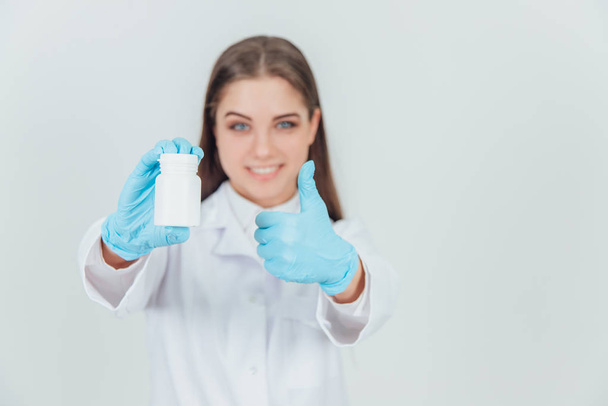 Blurred pretty young intern girl holding white bottle of pills, giving thumb up, showing her approval. Focus on the bottle on the forefront. - Foto, Imagem
