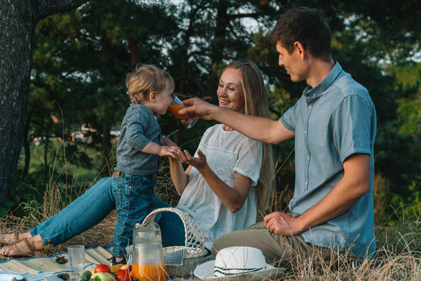 Mother and father with young son having picnic in park. The father gives the child to drink juice from a glass. Family leisure outdoors in the natural landscape - Foto, afbeelding