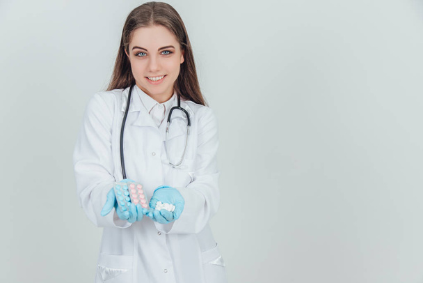 Adorable young nurse standing with one palm full of white pills. Two blisterpacks of colorful pills in another palm. - Foto, Bild