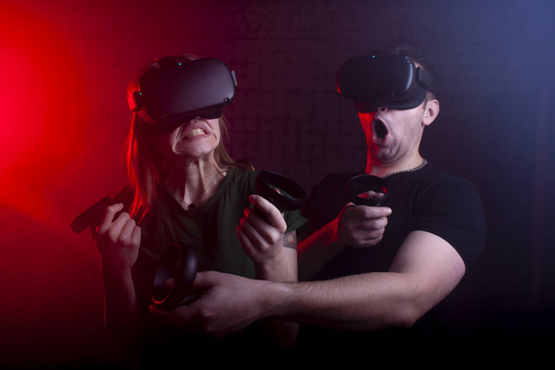 paar in moderne glazen van virtual reality spelen een shooter tegen een donkere neon achtergrond, een team van gamers in een spel met wapens - Foto, afbeelding