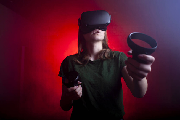 meisje gamer in moderne virtual reality bril speelt een shooter op een donker rood-blauwe achtergrond, een vrouw schiet met spel joysticks - Foto, afbeelding