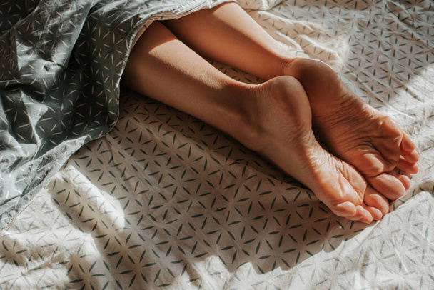 Feet of woman peeking out from under the covers. Girl alone sleep in bed. Wake up. Female legs. Happy morning in bedroom. Cozy and comfortable. Sunlight on bed linen. Pillow, blanket. Recovery, relax - Foto, Imagem