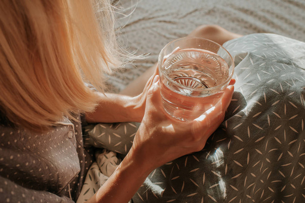 Woman in bed holding a glass of water in hand. Happy morning. Girl in pajamas. Healthy lifestyle, wellness. Proper nutrition. Drinking water. Morning with water. Sunlight on linens. Pillow, blanket - Фото, изображение