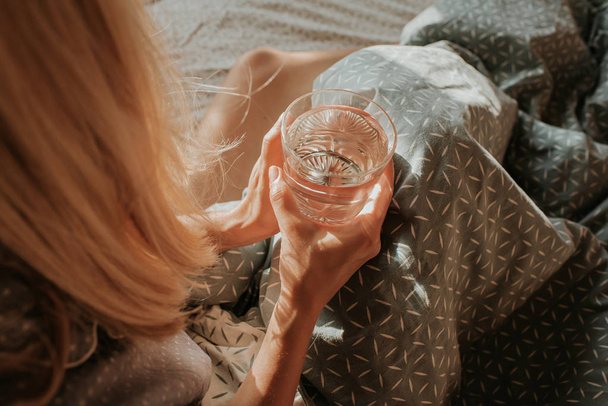 Woman in bed holding a glass of water in hand. Happy morning. Girl in pajamas. Healthy lifestyle, wellness. Proper nutrition. Drinking water. Morning with water. Sunlight on linens. Pillow, blanket - Foto, immagini