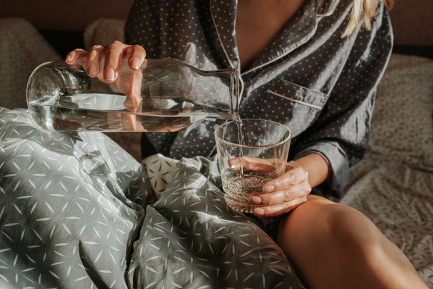 Woman in bed holding a glass of water in hand. Happy morning. Girl in pajamas. Healthy lifestyle, wellness. Proper nutrition. Drinking water. Morning with water. Sunlight on linens. Pillow, blanket - Valokuva, kuva