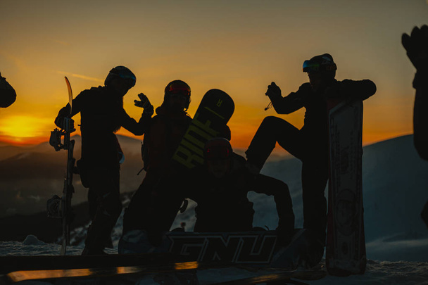 silhouettes of snowboarders in winter clothes with snowboards relaxing on hill in winter mountains during sunset    - Photo, Image