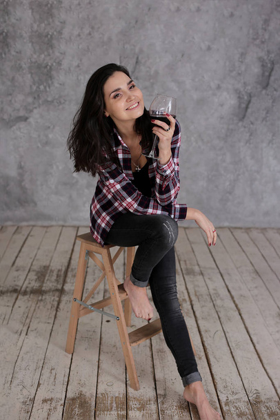 Krásná štíhlá dívka s krátkými tmavými vlasy v vínové kostkované košili se sklenkou červeného vína v podkroví šedý vnitřní styl - Fotografie, Obrázek