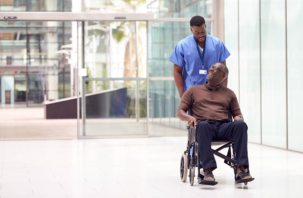 Male Nurse Wearing Scrubs Wheeling Patient In Wheelchair Through Lobby Of Modern Hospital Building - Фото, изображение