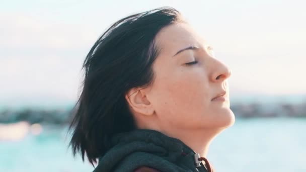 Profile of a natural brunette woman with no makeup enjoying the breeze and the sunshine with eyes closed near the sea. - Video, Çekim