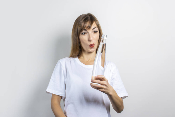 Young woman with a surprised face holds a bottle of clear water on a light background. Banner. Concept of thirst, heat, health and beauty care, water balance - Photo, Image