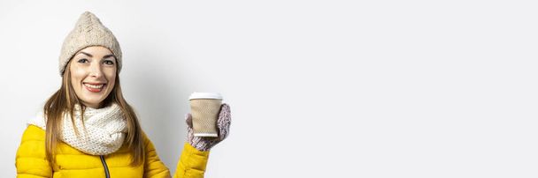 Junge Frau in gelber Jacke und Hut hält ein Glas Kaffee oder Tee vor hellem Hintergrund. Emotionales Lachen, Überraschung, Kuss. Konzeptwinter, Herbst, Kaffeehaus. Banner - Foto, Bild