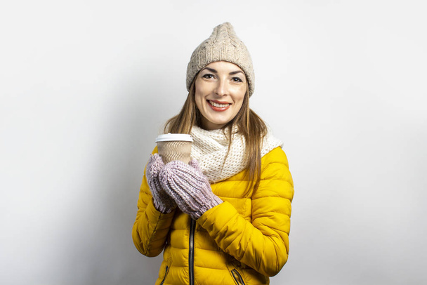 Mladá žena ve žlutém saku a klobouku drží sklenici kávy nebo čaje na světlém pozadí. Emoční smích, překvapení, polibek. Koncept zima, podzim, kavárna - Fotografie, Obrázek