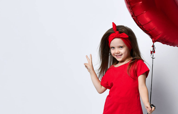 Small girl in red t-shirt and hair accessory standing, holding red balloon in hand and showing peace sign over white background. - Foto, afbeelding